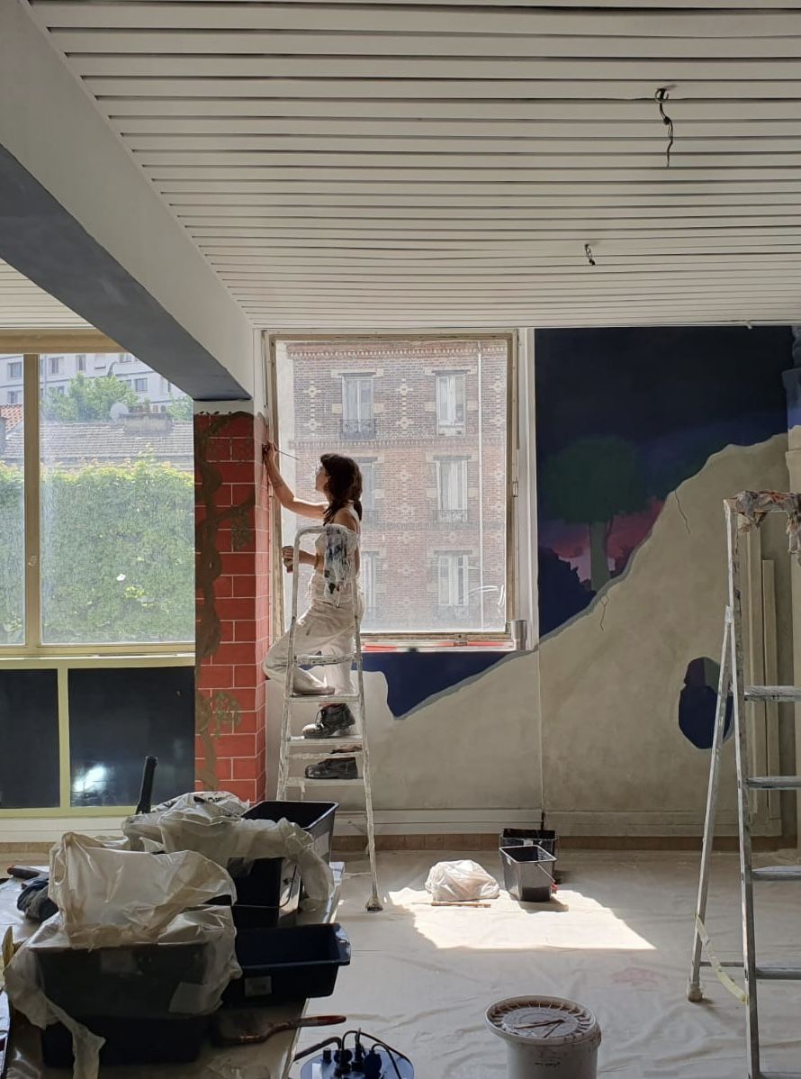 Félicie sur un escabeau peignant un pilier en fausses briques, à côté d'un grand décor mural de paysage