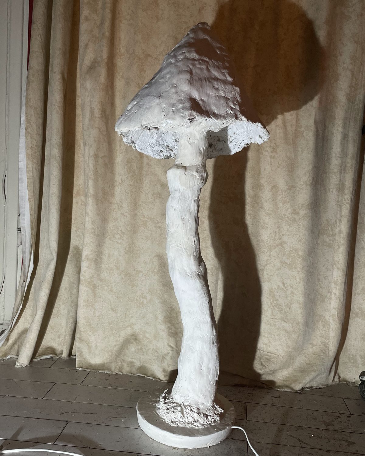 Luminaire sculpté en plâtre blanc en forme de champignon, au pied élancé, devant un rideau beige